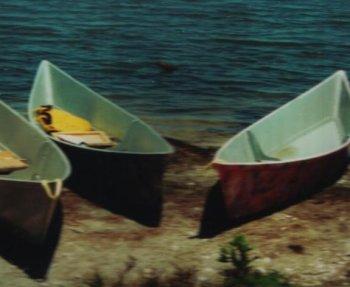 The image shows three canoes varying in color, resting on a sandy shore, close to a lake.