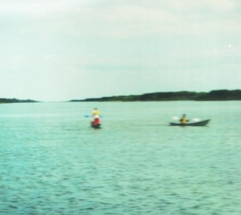 The image shows a wide, calm lake under a slightly overcast sky. Two student-built canoes are visible on the water.