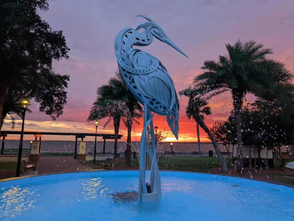 Estatua de garza y fuente del Parque Ferran en Eustis, FL, con un vibrante atardecer sobre el agua.
